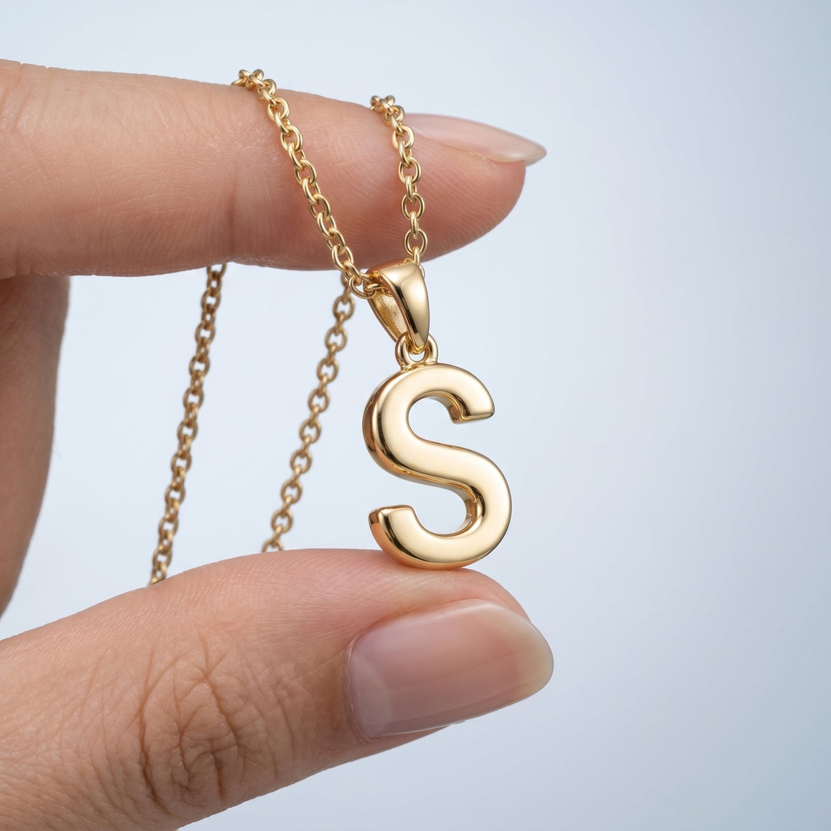 Collar con inicial de imprenta bañado en oro de 18K sostenido entre los dedos, para ver el tamaño real