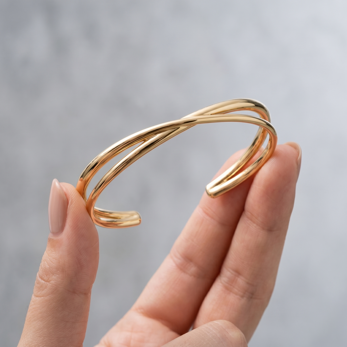 Brazalete rígido cruzado bañado en oro de 18K sostenido entre los dedos, para ver el tamaño real