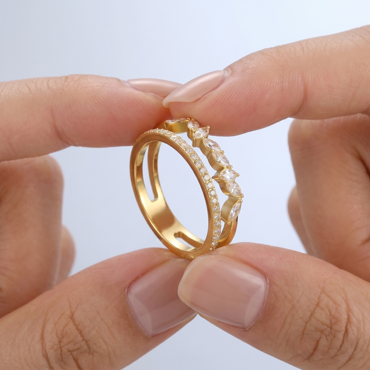 Anillo de doble banda pavé con cristales bañado en oro de 18K sostenido entre los dedos, para ver el tamaño real
