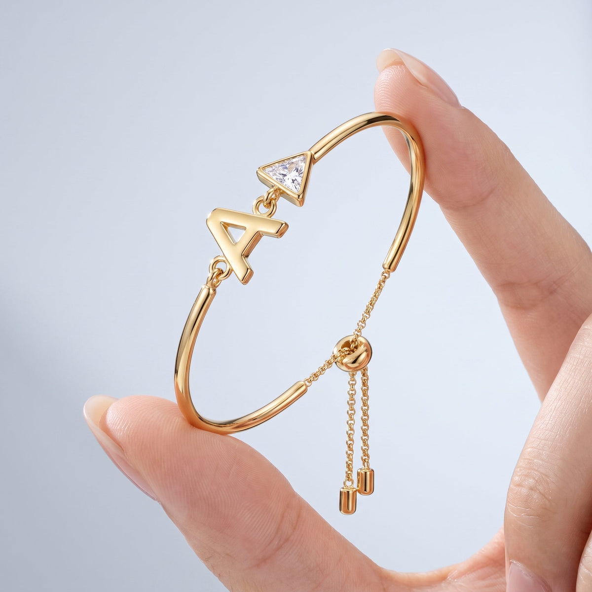Pulsera con inicial y cristal bañada en oro de 18K sostenida entre los dedos, para ver el tamaño real