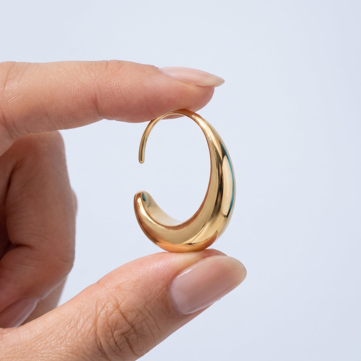 Pendientes ovalados pasantes bañados en oro de 18K sostenidos entre los dedos, para ver el tamaño real