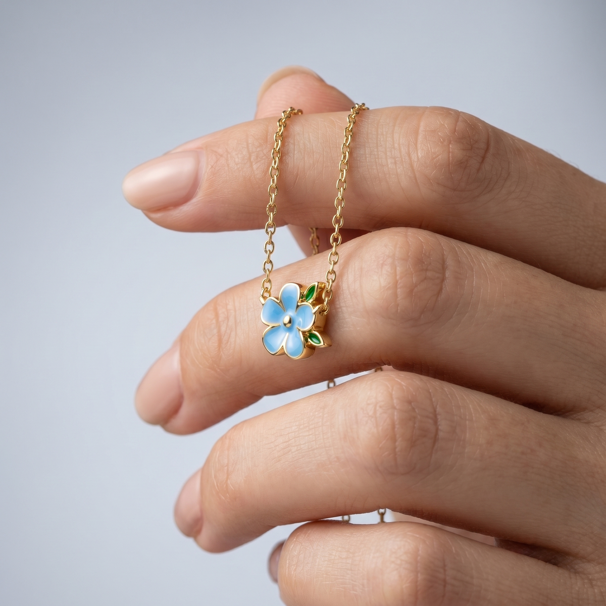 Collar con flor esmaltada azul bañado en oro de 18K sostenido entre los dedos, para ver el tamaño real
